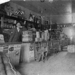 A black and white historical photograph of an early 20th-century general store. A man with a mustache, wearing a suit and hat, stands behind a wooden counter filled with various goods. Shelves behind him are stocked with bottles, tins, and other merchandise. The store is cluttered with wooden crates, barrels, and sacks. The ceiling is made of wood, and light streams in from a window or door on the right side.