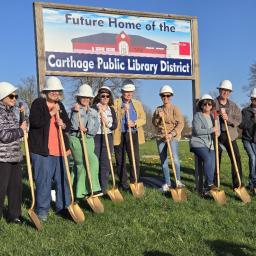 Photo of Library director & board members wearing hard hats and with gold shovels. Sign "Future home of the Carthage Public Library District"