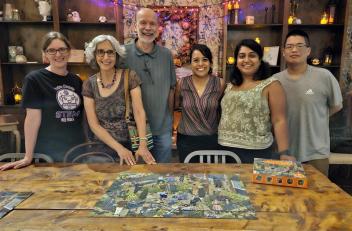 Six friends stand together smiling. In front of them is a table with a completed puzzle.