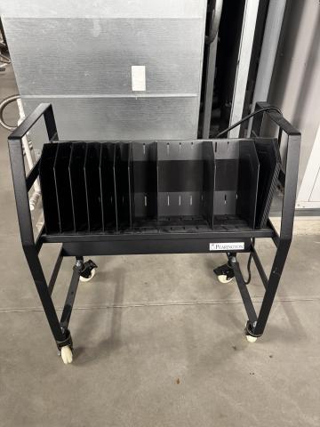 Black metal rolling storage cart with multiple vertical dividers creating slots for equipment that is charging. The cart sits on four caster wheels. 